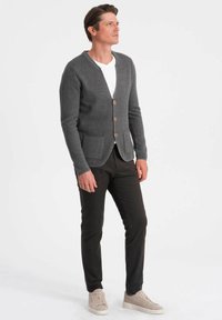 Grå stickad cardigan med V-ringning, träknappar, två framfickor, bärs över en vit T-shirt, ihop med mörka byxor och ljusa sneakers.