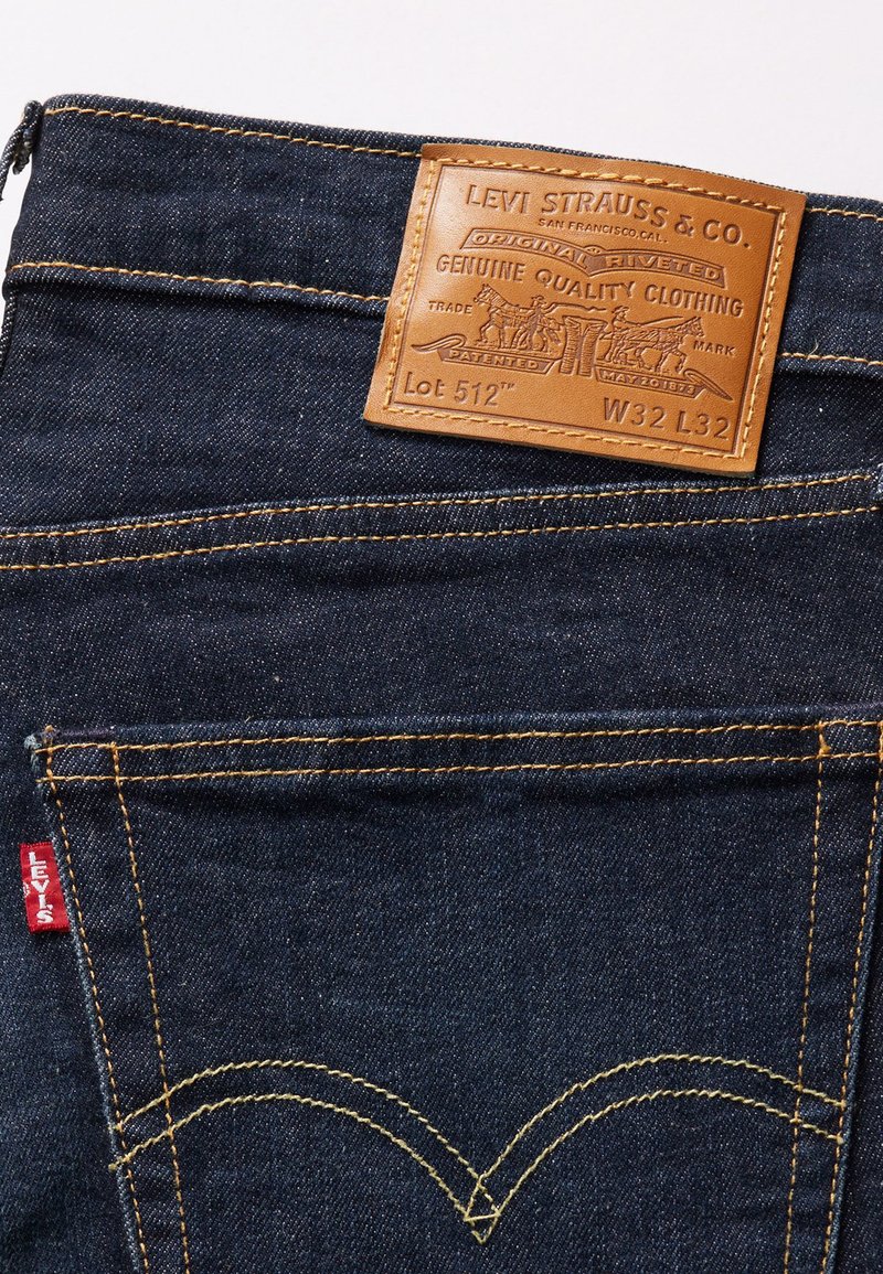 Jean en denim bleu foncé avec une étiquette en cuir embossée. Coutures jaunes et une étiquette rouge sur la poche arrière sont visibles.