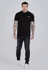 Trumpa juoda marškinėliai su trumpomis rankovėmis ir mažo dydžio logotipu ant krūtinės, derinami su juodais, dėvėtais džinsais ir baltais sportbačiais. Aiški, prigludusi silueto linija.