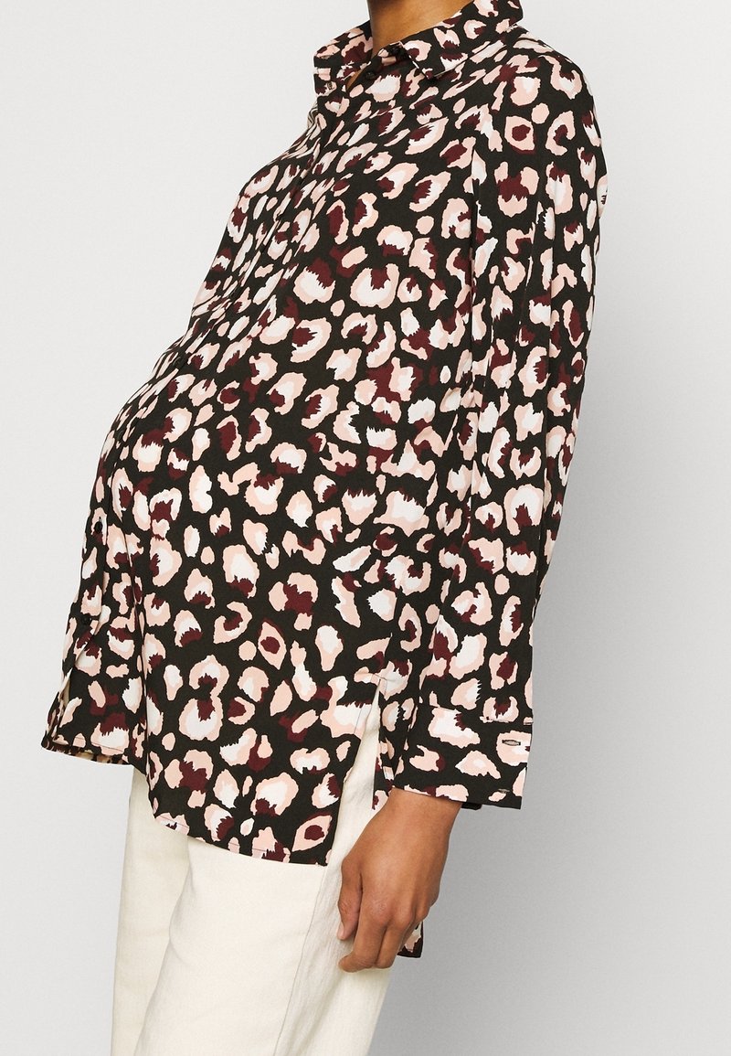 Femme portant une chemise ample à boutons avec un motif léopard noir, rose et bordeaux, et un pantalon blanc cassé.