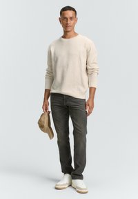 Beige Strickpullover mit langen Ärmeln, graue Jeans und weiße Sneakers mit beigen Akzenten; hält eine hellbeige Baseballkappe.