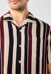 Camisa de manga corta con cuello, con rayas verticales en burdeos, crema y azul marino. Incluye un bolsillo en el pecho y cierre de botones.