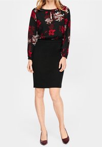 Femme portant une jupe crayon noire et un chemisier noir à fleurs rouges et blanches, assortis à des escarpins pointus rouge foncé.