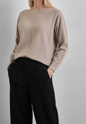 Vrouw draagt een beige trui met lange mouwen en een centrale naad, gecombineerd met zwarte, hooggesneden broek met zijknopen en handen in de zakken.