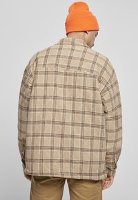 Beżowa koszula-kurtka w kratę z pikowanym wzorem, z brązowymi elementami w kratkę i kołnierzykiem zapinanym na guziki. Noszona z pomarańczową czapką beanie.