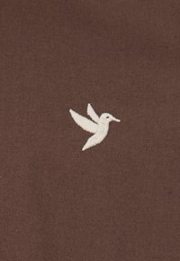 Tejido marrón con un acabado texturizado que presenta un diseño bordado de colibrí blanco, mostrando alas detalladas y un cuerpo esbelto.