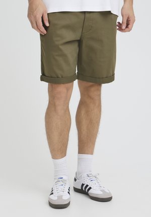 Mand iført olivengrønne shorts med opsmøg, hvide crew-sokker og hvide sneakers med sorte striber, som står op ad en ensfarvet baggrund.