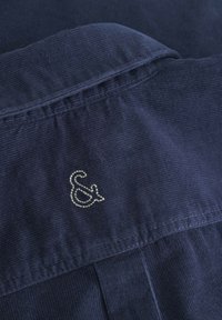 Camicia in corda blu navy con tessuto testurizzato, caratterizzata da un logo decorativo argento "&" ricamato sul colletto posteriore.