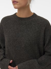 Persona con un maglione di lana grigio scuro, con colletto a costine e maniche lunghe, che tiene parzialmente le mani vicino alla vita, indossando orecchini con perle a goccia.