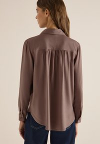 Femme aux longs cheveux bruns portant un chemisier ample taupe avec un empiècement froncé dans le dos et un jean bleu foncé, vue de profil sur fond beige.