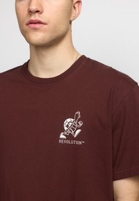 Bordeaux katoenen t-shirt met een witte afbeelding van een schildpad en de tekst "REVOLUTION™" op de linkerkant van de borst, met een ronde hals.