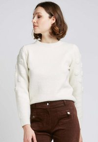 Pull en tricot blanc avec un col rond et des détails en relief sur les manches, associé à un pantalon en velours côtelé marron.
