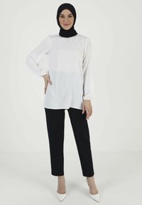 Langärmlige Bluse in Weiß, mit geradem Schnitt und elastischen Bündchen, kombiniert mit schwarzen geraden Hosen und weißen spitzen Absätzen.