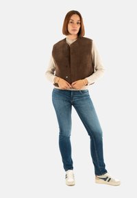 Gilet in pile marrone con chiusura a bottoni, indossato sopra un maglione chiaro, abbinato a jeans blu e sneakers beige con accenti scuri.