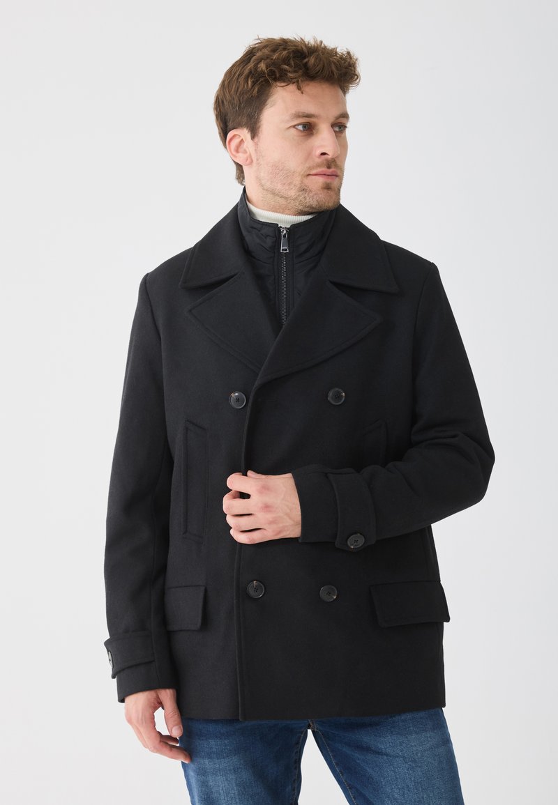 Schwarzer doppelt gefütterter Mantel mit breiten Revers, Seitentaschen und Knopfakzenten; drüber eine innere Jacke mit Reißverschluss; der Stoff wirkt glatt.