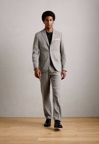 Hombre caminando hacia adelante con un traje gris claro, camisa negra y zapatos negros contra una pared beige lisa y un suelo de madera.