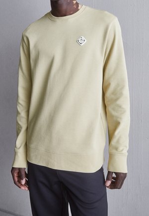 Person iført en bleggul langærmet sweatshirt med et lille diamantformet logo på venstre bryst og mørke bukser foran en grå væg.