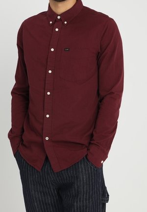 Mann trägt ein geknöpftes bordeauxrotes Langarmhemd mit Brusttasche und dunkle Nadelstreifenhose, die Hände in den Taschen.