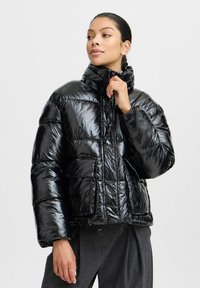 Veste rembourrée noire brillante avec un col montant, fermeture éclair et poignets élastiques. Présente un design matelassé et deux poches latérales.
