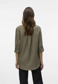 Olijfgroene button-up shirt met opgerolde mouwen, rugpasdetailing en een gebogen zoom, gemaakt van een gestructureerde stof. Gecombineerd met een donkere broek.