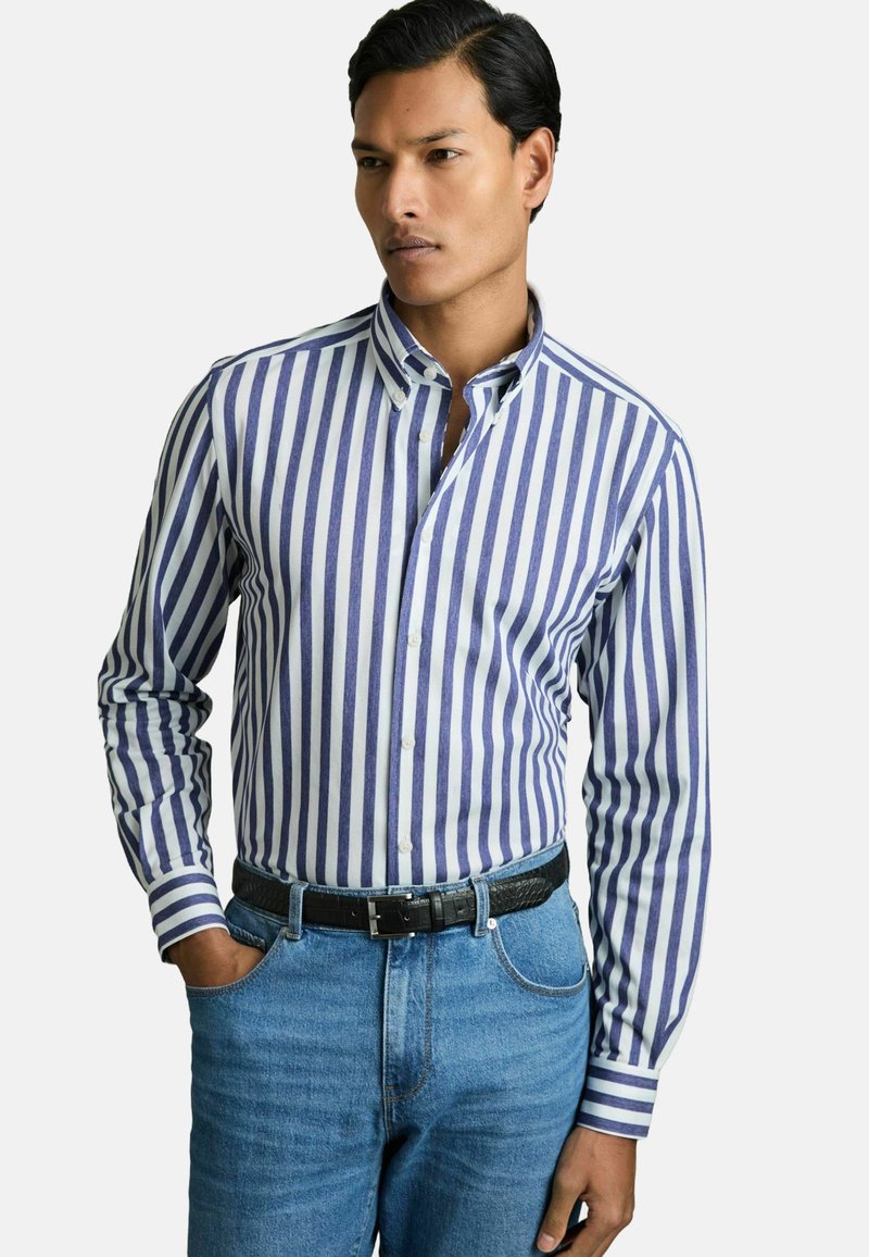 Camisa de botones a rayas en blanco y púrpura, con un cuello clásico y mangas largas, combinada con jeans de mezclilla azules y un cinturón negro.