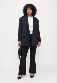 Svart oversized blazer med to frontlommer, parret med en hvit t-skjorte og svarte utsvungne bukser. Tilbehør inkluderer en svart perleveske.