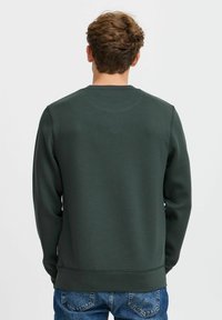 Mørkegrønn genser laget av mykt materiale, med klassisk rund hals, lange ermer og ribbad kant. Brukt sammen med blå denim jeans.