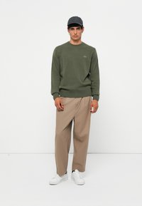 Green knitted sweater with ribbed cuffs and hem, paired with loose beige trousers and white sneakers, worn with a dark cap.