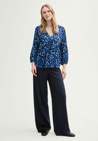 Blusa blu con un motivo a pois, scollatura a V e maniche a palloncino, abbinata a pantaloni blu navy a gamba larga e scarpe nere. Tessuto dalla consistenza liscia.