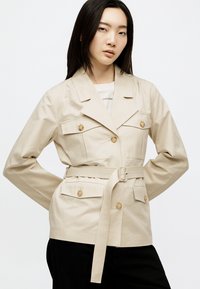 Γυναίκα με μακριά μαύρα μαλλιά που φοράει μπεζ σακάκι με ζώνη και κουμπωτές τσέπες πάνω από λευκό τοπ και μαύρο παντελόνι, στέκεται με τα χέρια στη μέση.