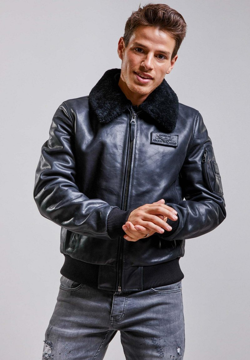 Blouson en cuir noir avec un col en fourrure, fermeture éclair sur le devant, poignets côtelés et un écusson sur la poitrine. Porté avec un jean gris déchiré.