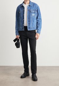 Giacca di jeans blu chiaro con chiusura a bottoni, tasche sul petto e patch con logo. Pantaloni neri e scarpe nere, tiene una piccola borsa nera.