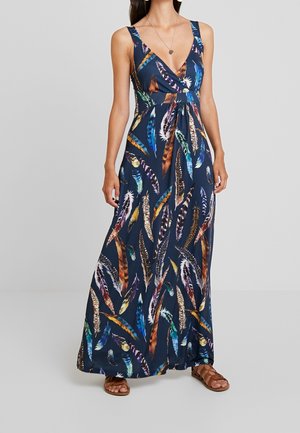 Vrouw draagt een mouwloze marineblauwe maxi-jurk met meerkleurige veerpatronen en bruine platte sandalen, staand tegen een effen grijze achtergrond.