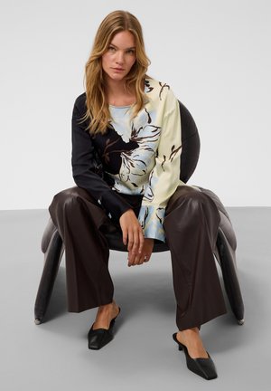 Femme aux longs cheveux blonds assise sur une chaise moderne noire, portant un haut à motif floral, un pantalon large et sombre, et des chaussures noires à talons bas.