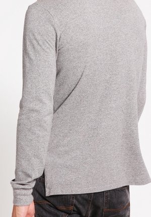 Mężczyzna ubrany w jasnoszary sweter z długim rękawem i czarne jeansy, przedstawiony od tyłu na jednolitym białym tle.