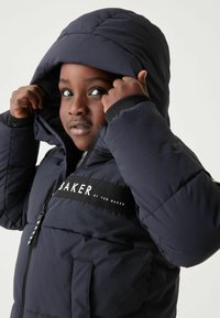 Doudoune navy avec capuche, présentant une bande logo noire sur la poitrine, fermeture éclair et texture matelassée.