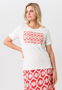 Witte T-shirt met korte mouwen en een rood geometrisch patroon. Soepele stof; ontspannen pasvorm. Gecombineerd met een rok met een vergelijkbaar rood ontwerp.