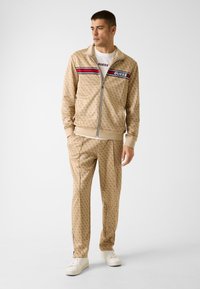 Tuta beige con motivo geometrico, giacca con zip e pantaloni abbinati. Presenta strisce rosse e bianche a contrasto; indossata con scarpe da ginnastica bianche.