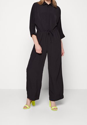 Nainen, jolla on päällä musta pitkähihaisen haalarin vyötärösoljella ja neonkeltaiset avokkaat, seisoo yksinkertaista vaaleaa taustaa vasten.