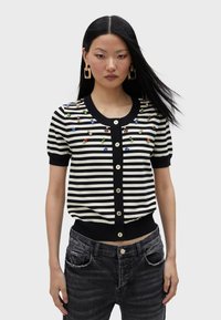 Crno-bijeli prugasti kardigan kratkih rukava sa ukrasima od dragulja i velikim dugmadima. Pleteni materijal s uskim strukom.