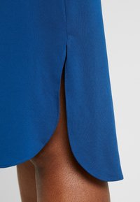 Blue fabric dress with a side slit revealing dark skin on the upper thigh against a neutral background.