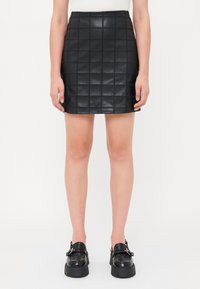 Jupe en simili cuir matelassé noire avec une coupe ajustée, présentant un motif en carrés et une taille. Associée à des mocassins noirs épais.