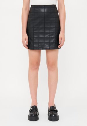 Svart quiltet fuskeskinnskjørt med en tettsittende fasong, med et firkantet mønster og en midjebånd. Kombinert med chunky svarte loafers.