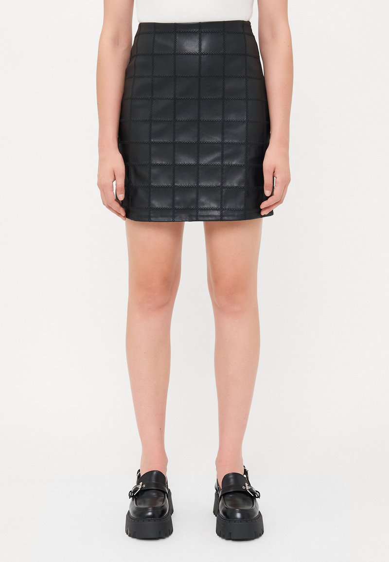 Jupe en simili cuir matelassé noire avec une coupe ajustée, présentant un motif en carrés et une taille. Associée à des mocassins noirs épais.