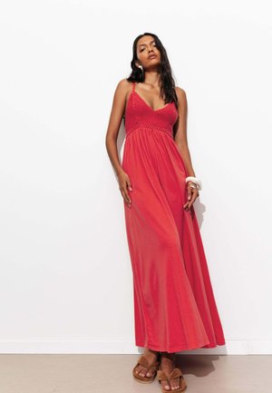 Femme aux longs cheveux foncés portant une robe longue rouge sans manches et des sandales marron, debout contre un mur blanc.