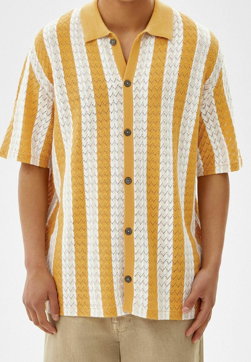 Kortärmad skjorta med knappar som har ett zigzag-strickmönster i gult och vitt, med en kontrasterande krage och textilstruktur.