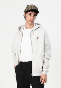 Lys grå zip-up hettegenser med rosebroderi på venstre bryst, kombinert med en hvit t-skjorte og svarte bukser, brukt sammen med en rutete caps.