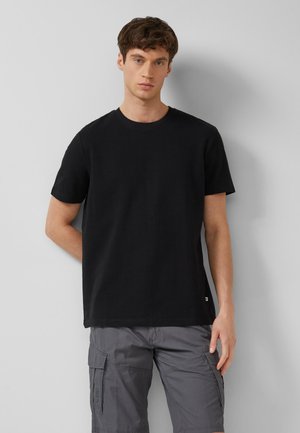 Young man with short brown hair wearing a plain black t-shirt and gray cargo shorts standing against a light gray wall.