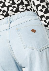 Jeans de denim azul claro com um acabamento texturizado, apresentando um patch de logo em couro castanho e dois bolsos traseiros, combinados com uma blusa estampada em preto e branco.
