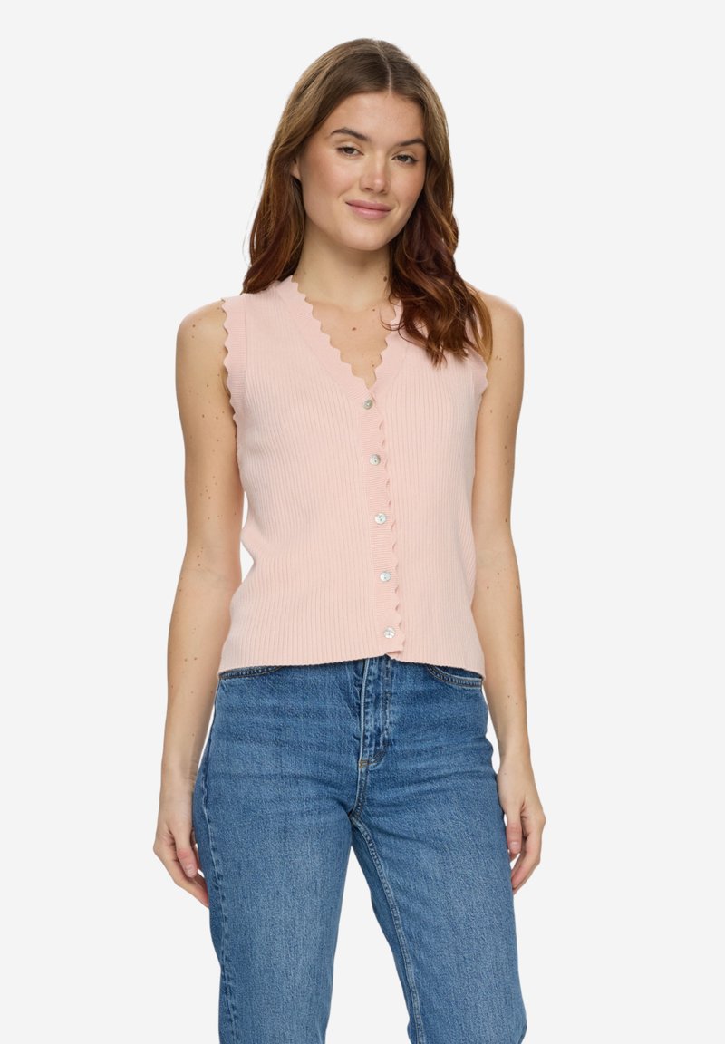 Femme aux cheveux bruns portant un haut côtelé sans manches rose clair à boutons et un jean taille haute bleu, debout devant un fond blanc.